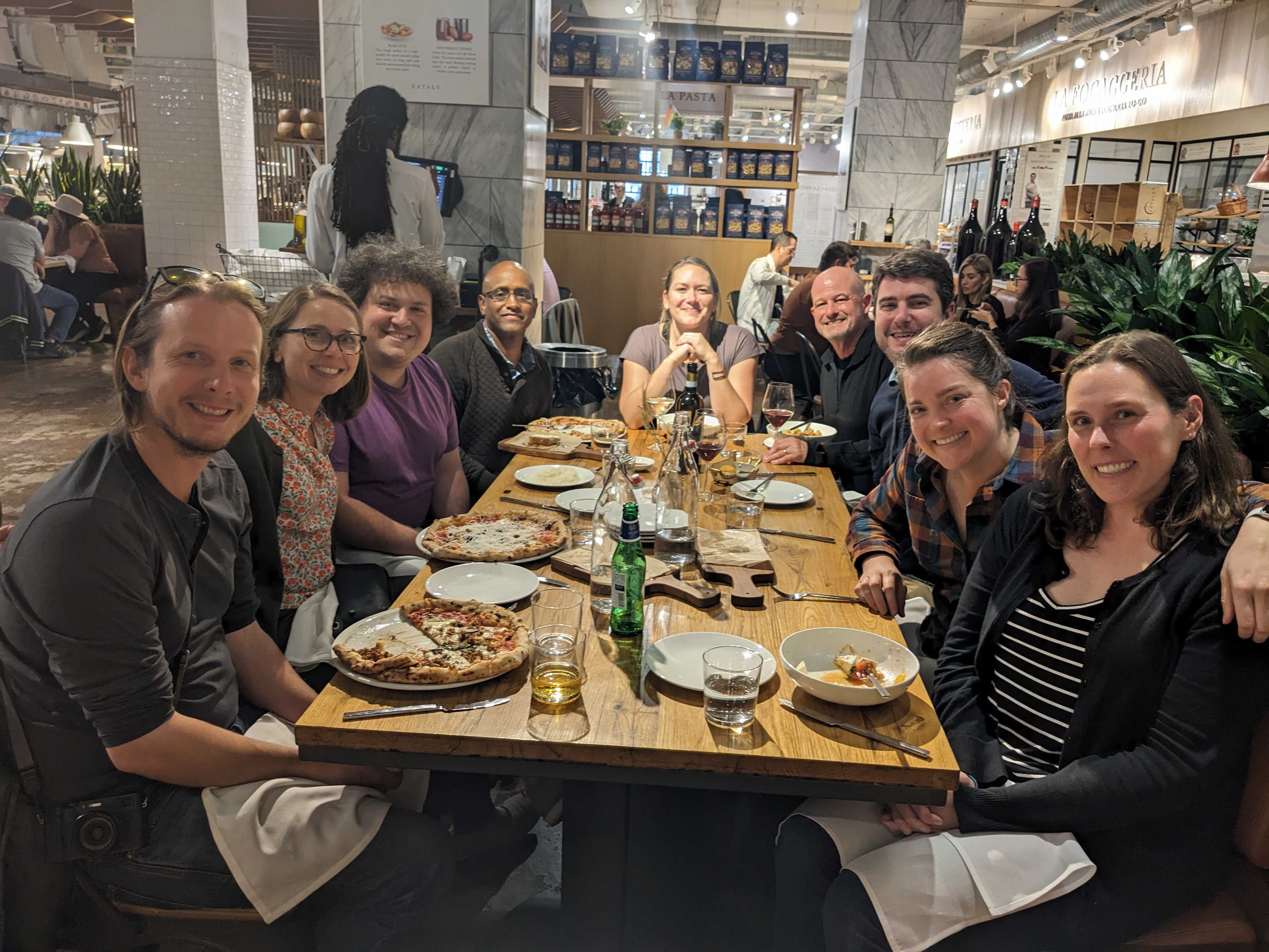 photo of 9 people seated around a rectangular restaurant table with restaurant interior in background. Some have pizzas on their plates.